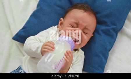 Hispanischer Junge, der weint und eine Milchflasche an den Mund hält, auf einem blauen Kissen in einer Futterszene im Studio, mit sichtbaren Tränen und geballter Hand; Hunge Stockfoto