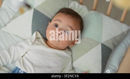 Der hispanische Junge, der in einem hölzernen Kinderbett auf einem gemusterten Kissen liegt, blickt mit weit offenen Augen und einem sichtbaren Gesicht auf die Kamera im Gebäude. Neugier Stockfoto