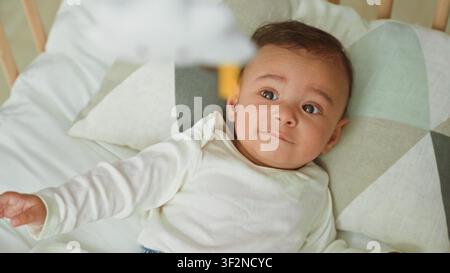Babyjunge liegt in der Wiege auf geometrischem Kissen und greift nach dem hängenden Mobile über ihm, Gebäude; Neugier verbindet Elternschaft. Stockfoto