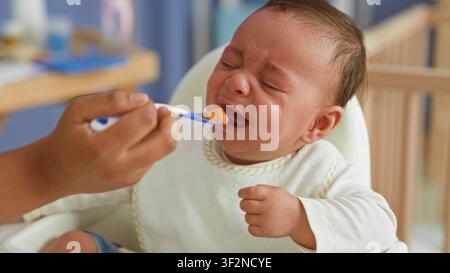 Hispanisches Baby in einem Hochstuhl, das mit dem Löffel gefüttert wird, Essen im Mund und mit verkniffenem Gesicht greifend, weinende Geste in einem Gebäude; Frustrationsverweigerung. Stockfoto