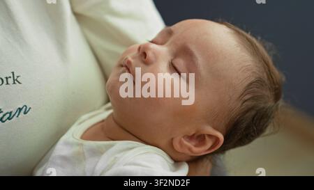 Hispanischer Junge, der auf dem Arm des Erwachsenen schläft, während die Hand das Gesicht sanft mit dem Tuch im Gebäude abwischt; sanfte Pflege beruhigt. Stockfoto