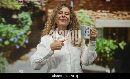 Die hispanische Frau im mittleren Alter zeigt mit dem Finger auf den Becher und liest: ich bin der Boss in einem Gebäude, lächelt und hält den Becher in der rechten Hand; Selbstvertrauen stärken Stockfoto