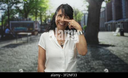 Eine ältere Frau zeigt beim Lächeln mit dem Finger an die Nase, trägt weiße Bluse und Anhänger auf einer Kopfsteinpflasterstraße. Stockfoto