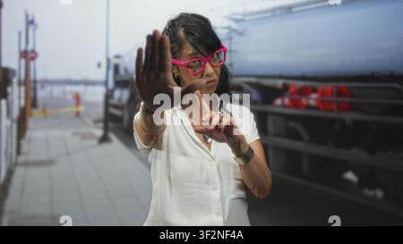 Frau Senior hispanic trägt eine rosafarbene Brille, hält die Handfläche hoch und zeigt mit dem Zeigefinger, um den Verkehr auf der Straße in der Nähe von LKW und Geländer zu stoppen; Warnbehörde. Stockfoto