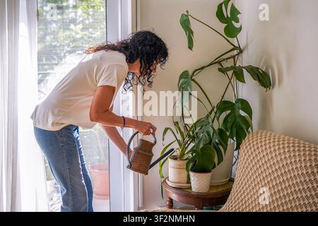 Mittlere erwachsene Frau lehnt sich nach vorne, während sie Zimmerpflanzen mit einem Vintage-Gießkann gießt Stockfoto