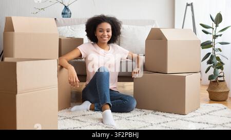 Lächelndes afroamerikanisches Mädchen, das auf dem Boden sitzt und von vielen Kisten umgeben ist Stockfoto
