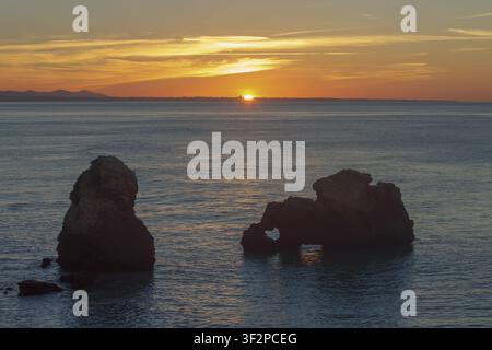 Die Sonne geht am Horizont unter und wirft Licht auf das Wasser. Zwei große Felsen heben sich im Vordergrund ab. Der Himmel hat Schattierungen von Orange und Rosa, während der Tag Stockfoto