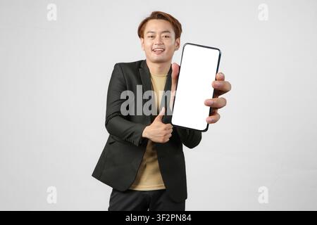 Porträt eines glücklichen asiatischen Geschäftsmannes im Anzug, der ein Mobiltelefon hält und einen leeren Bildschirm zeigt, während er auf isoliertem Hintergrund einen Daumen hoch zeigt Stockfoto