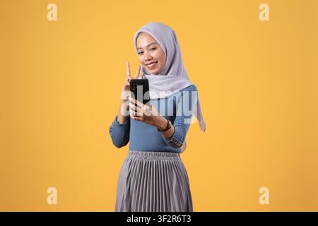 glückliche asiatische indonesische muslimische Frau, die ein Smartphone hält und auf isoliertem gelbem Hintergrund den Siegesfinger zeigt Stockfoto