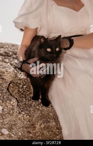 Katzenfrau mit Handschuhen hält eine flauschige schwarze Katze auf texturiertem Stein, während sie ein elegantes weißes Leinenkleid trägt Stockfoto