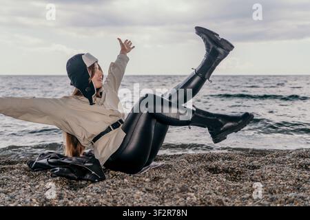 Frau Strandglück spielerische Frau in Fliegerhut und Stiefeln sitzt am Kieselufer mit angehobenen Beinen an einem Wintertag Stockfoto