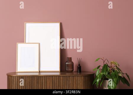 Zwei leere Holzbildrahmen auf einem gerillten Holzschrank mit einer Zimmerpflanze und einem Diffusor vor einer rosa Wand, die Anordnung ist vor einem festen Hintergrund platziert Stockfoto