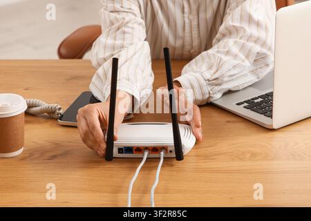 Person, die Ethernet-Kabel an einen weißen drahtlosen Router auf einem Holzschreibtisch mit einem Laptop und einer Kaffeetasse anschließt Stockfoto