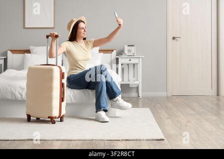 Junge Frau mit Strohhut, die ein Selfie mit einem Smartphone macht, während sie auf einem Bett neben einem Koffer in einem Hotelzimmer sitzt, ein Koffer steht neben ihr, Stockfoto