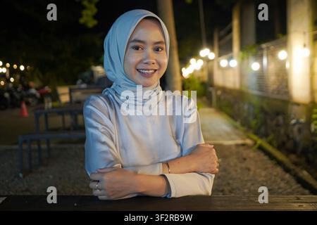 Fröhliche asiatische muslimische Frau mit Hijab, die in die Kamera lächelt, während sie im Freien an einem Nachtmarkt-Food-Court sitzt Stockfoto