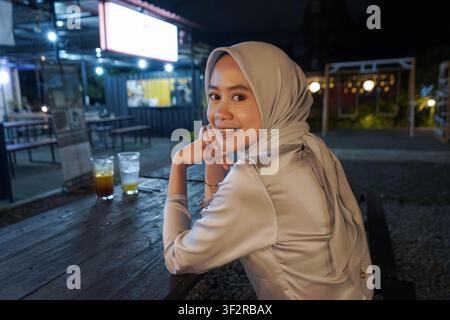 Fröhliche asiatische muslimische Frau mit Hijab, die in die Kamera lächelt, während sie im Freien an einem Nachtmarkt-Food-Court sitzt Stockfoto