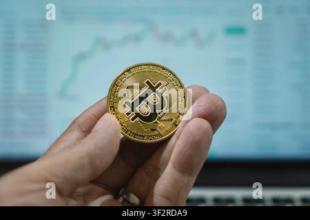 Nahaufnahme einer Männerhand, die Bitcoin hält, mit verschwommenem Bildschirm, der ein Diagramm im Hintergrund anzeigt Stockfoto