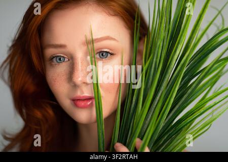 Ginger-Haarige Junge Frau, Die Die Kamera Durch Die Pflanze Sieht, Grauer Hintergrund Stockfoto