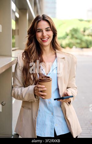 Selbstbewusste junge Geschäftsfrau steht im Freien, hält einen wiederverwendbaren Kaffeebecher und ein Smartphone und präsentiert Professionalität und Erfolg Stockfoto