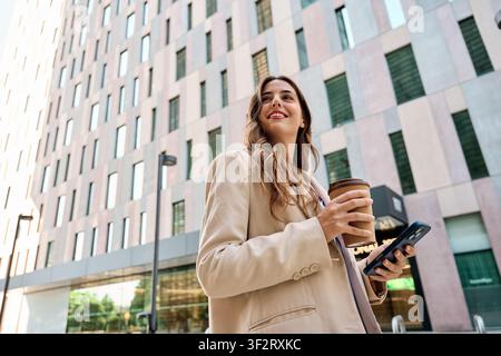 Selbstbewusste Geschäftsfrau geht vor Bürogebäude, lächelt und schaut weg, während sie eine Kaffeetasse und ein Smartphone hält Stockfoto