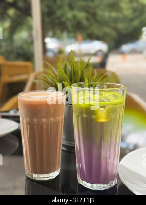 Schokoladenmilchgetränk und farbenfrohes geschichtetes Eismatcha-Latte, serviert in hohen Gläsern auf einem Außentisch eines Cafés. Moderne Sommergetränkekomposition mit Stockfoto