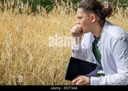 Nachdenklicher Bauer hockt in hohem Weizenfeld, Hand am Kinn, während er Tablet und Ordner hält. Mann denkt über die Analyse der Erntequalität nach während der Landwirtschaft Stockfoto