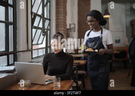 Kellnerin serviert ein Croissant an eine Frau, die an einem Laptop arbeitet Stockfoto