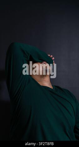 Dramatisches Porträt eines erwachsenen indonesischen Mannes in einem dunklen grünen Hemd, der sich zurücklehnt und die Augen mit einem Arm bedeckt, vor einem schwarzen Hintergrund. Stockfoto