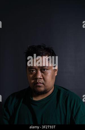 Nahaufnahme eines ernsten erwachsenen indonesischen Mannes in einem dunkelgrünen Hemd, der direkt in die Kamera blickt vor einem schroffen schwarzen Hintergrund. Der i Stockfoto