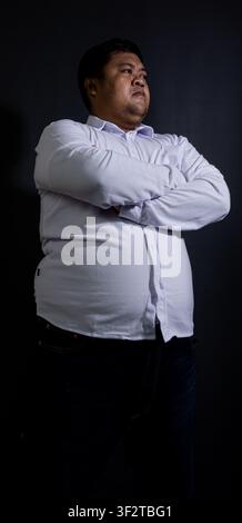 Erwachsener asiatischer Mann steht mit verschränkten Armen vor einem einfarbigen schwarzen Hintergrund, trägt ein langärmeliges weißes Hemd mit Knopfleiste und dunkelblaue Denimjeans Stockfoto