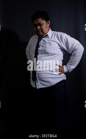 Übergewichtiger erwachsener asiatischer Mann, der mit den Händen auf den Hüften gegen einen dunklen schwarzen Hintergrund steht, trägt ein weißes langärmeliges Hemd, dunkelblaue Jeans, Stockfoto