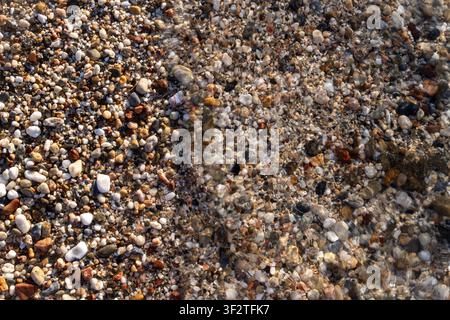 Transparente Meereswasserwelle spült über nasse mehrfarbige kleine Steine und Sand an der Küste während eines sonnigen Tages, Nahaufnahme. Stockfoto