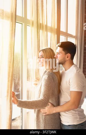 Junger Mann und Frau in der Liebe umarmen sich am sonnigen Tag in der Nähe von Fenster zu Hause, unbeschriftet Stockfoto