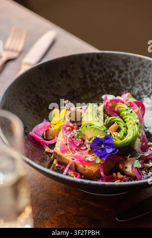 Nahaufnahme eines handwerklich zubereiteten Avocado-Toasts, gestylt mit einer Avocado-Rose, eingelegten roten Zwiebeln und bunten essbaren Blumen in einer dunklen, strukturierten Schüssel. Stockfoto