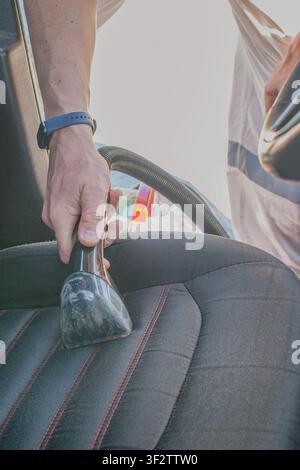 Nahaufnahme eines männlichen Detailers, der eine professionelle Saugdüse verwendet, um nassen Schmutz von einem schwarzen Textil-Autositzkissen in einer Autowerkstatt zu entfernen Stockfoto