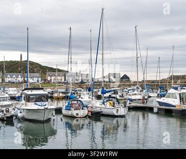 Boote ruhen im stillen Wasser im Hafen von Findochty an einem grauen Tag in Moray, Schottland, wo Häuser nahe der steinigen Küste liegen und hohe Masten emporragen Stockfoto