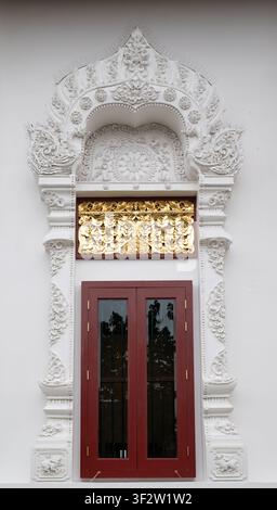 Chiang Mai, Thailand - 20. Juli 2026: Weißer Stuckrahmen eines Tempelfensters mit goldener Naga-Schnitzerei und roten Holzläden an einem buddhistischen Tempel Stockfoto