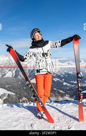 Lächelnde Skifahrerin genießt die Sonne, während sie rote Skier auf einem schneebedeckten Alpen-Gipfel hält. Majestätisches Bergpanorama, blauer Himmel, Wintersport, Freiheit Stockfoto