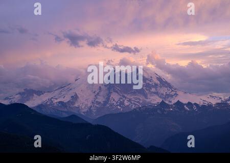 Blick auf den Mount Rainier bei Sonnenuntergang mit schneebedeckten Gipfeln, lila und rosa Wolken und dunklen bewaldeten Bergen im Vordergrund in Pierce County, Washin Stockfoto