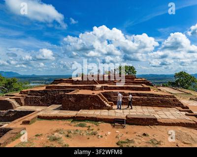 Luftaufnahme der antiken Ruinen des Sigiriya-Löwenfelsen, mit Steinfundamenten, Ziegelbauten und Touristen auf dem Gipfel unter einem blauen Himmel in Stockfoto
