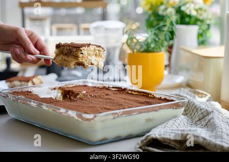 Eine Tiramisu-Scheibe wird mit einem Spatel aus einer Glasschale mit Kakaotopping auf dem Küchentisch serviert. Hausgemachtes Dessert und süße Speisenportionierung Stockfoto