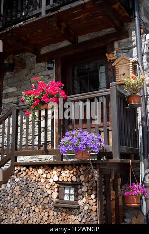 Ein rustikaler Holzbalkon geschmückt mit lebhaften rosa und lila Blumen über einem ordentlich gestapelten Holzstapel in einer sonnigen Bergdorflandschaft. Stockfoto
