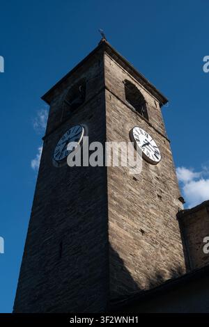 Eine niedrig angelegte Aufnahme eines alten steinernen Glockenturms mit zwei Uhren vor einem tiefblauen Himmel. Das Sonnenlicht betont die Textur des antiken Mauerwerks. Stockfoto