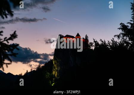 Niedrigwinkelaufnahme von Bled Castle, das auf einer felsigen Klippe über dem Bledsee in Slowenien thront, unter einem dramatischen dunklen Himmel nach Sonnenuntergang, wobei seine mittelalterliche Architektur betont wird Stockfoto