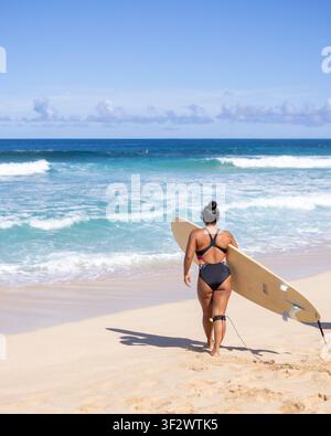 EHUKAI BEACH, NORTH SHORE, HAWAII - 11. FEBRUAR 2022: Eine Frau trägt ein Longboard in Richtung einer sauberen Welle der North Shore, von hinten im Sand gesehen. Stockfoto