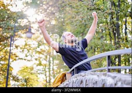 Mann hebt die Fäuste zum Sieg, während er auf einer Parkbrücke im Herbstsonnenlicht steht. Energetische Outdoorszene für Erfolg, Leistung, Motivation, Freiheit oder Lifestyle-Werbung Stockfoto