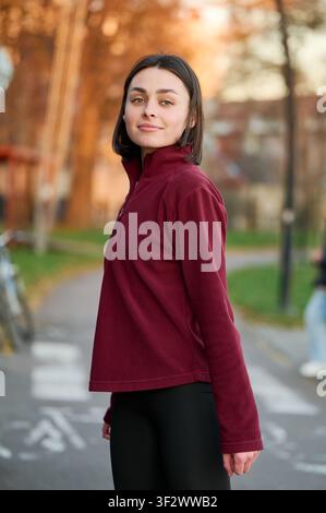Junge Frau steht auf einem städtischen Pfad in burgunderrotem Fleece, Nahaufnahme mit ruhigem Ausdruck im warmen Abendlicht. Nützlich für Lifestyle-, Mode-, Wellness- oder Outdoor-Redaktionsthemen Stockfoto