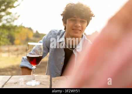 Junger Mann lächelt sanft am Tisch. Rotwein glüht im Glas neben ihm. Sonnenlicht taucht die Szene in warmes Licht. Er trägt ein lässiges Denimhemd und ein dunkles T-Shirt. Sett Stockfoto