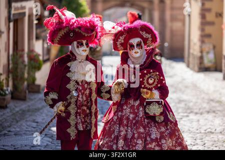 Riquewihr, Frankreich - 04. Juli 2026: Blick auf zwei Personen, die in aufwendigen venezianischen Karnevalskostümen in Rot und Gold gekleidet sind und auf einem historischen Kopfsteinpflaster stehen Stockfoto