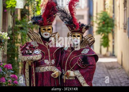 Riquewihr, Frankreich - 04. Juli 2026: Blick auf zwei Personen, die aufwendige rote und goldene venezianische Karnevalskostüme und Masken tragen, während sie in einem historischen Stockfoto